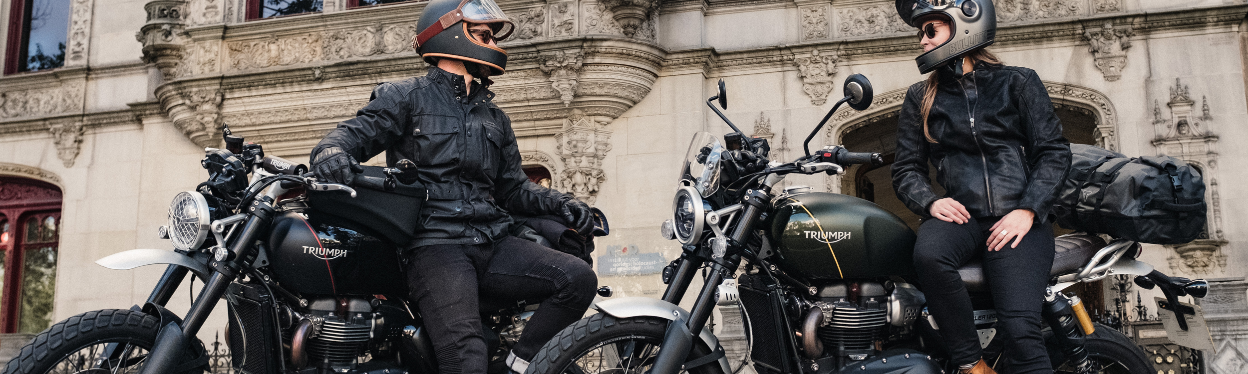 Two motorcycle riders sitting on their bikes and talking in front of historic building.