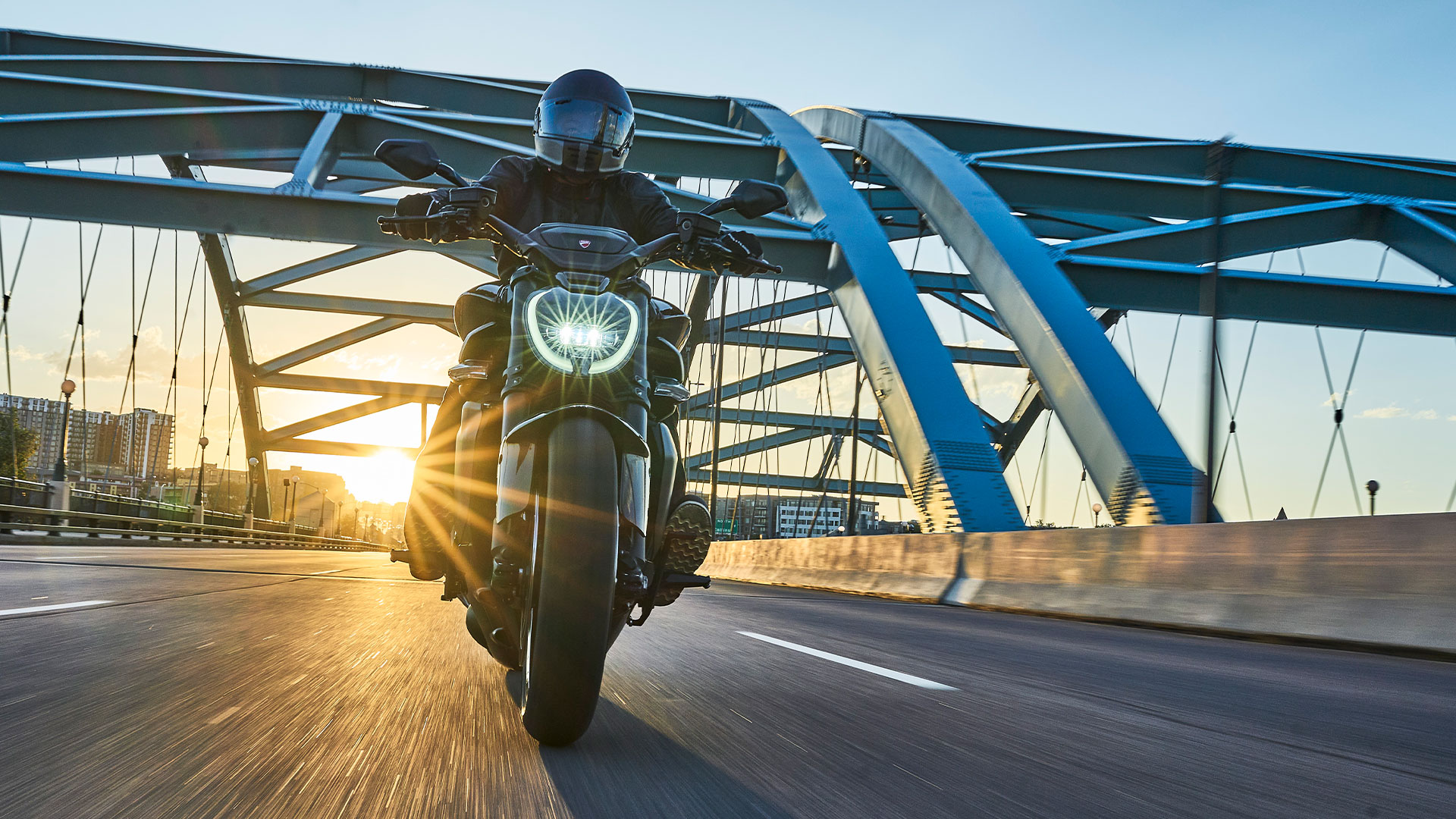 Motorcyclist riding a Ducati Diavel across a steel bridge at sunset